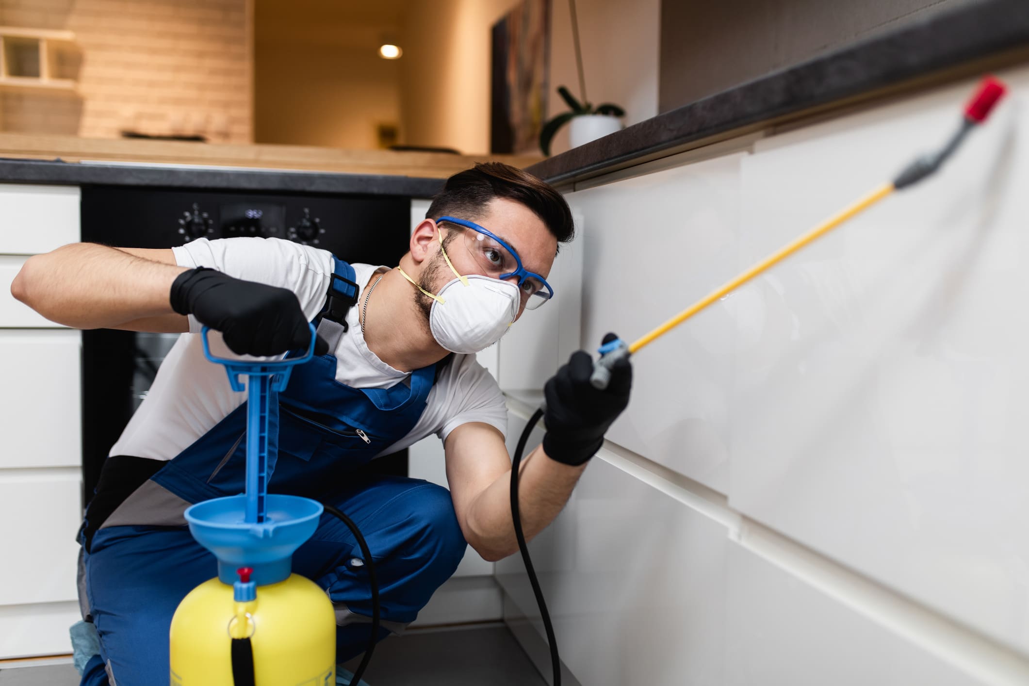 Professional exterminator in protective workwear spraying pesticide in apartment kitchen