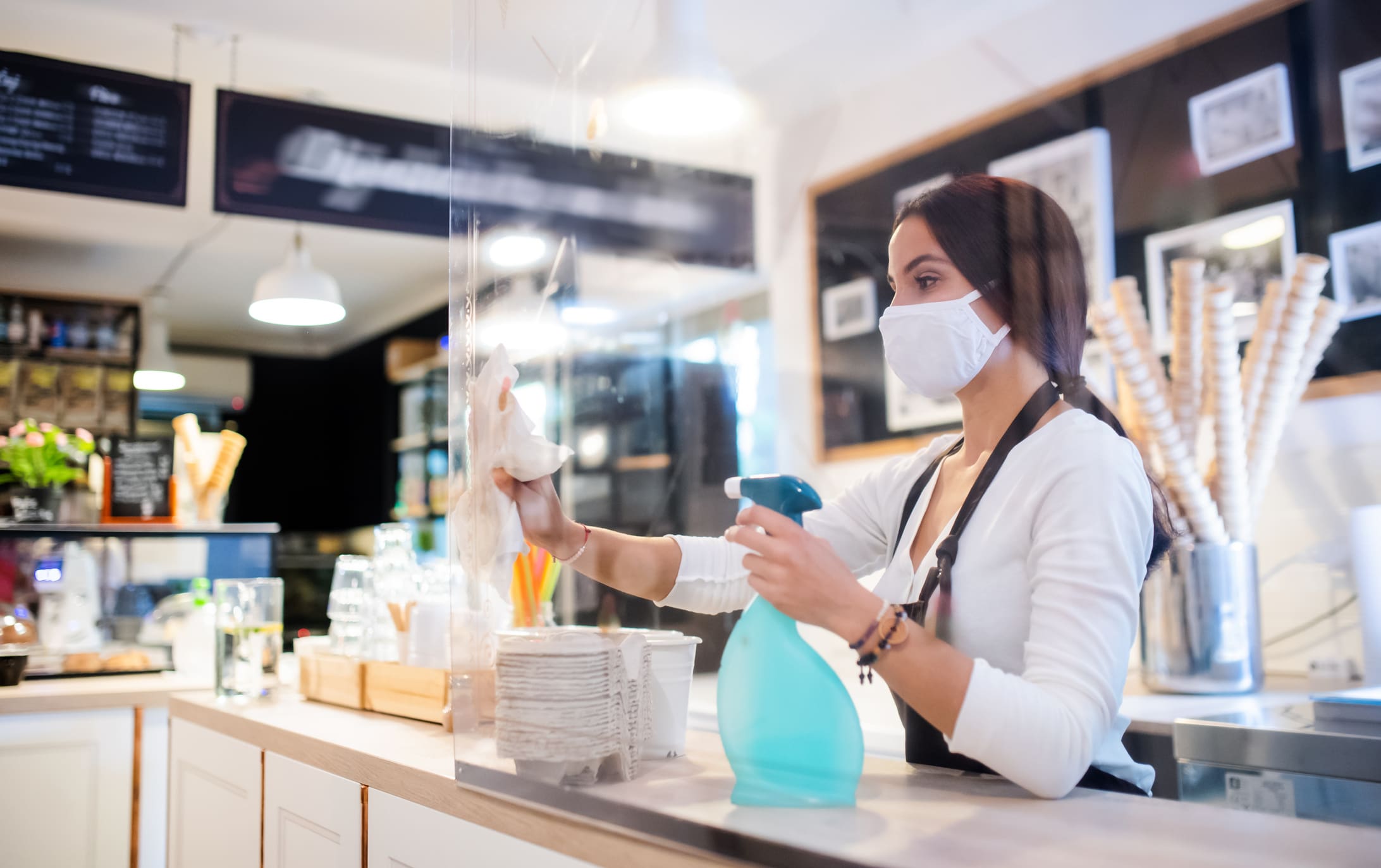 Portrait of young woman with face mask working indoors in café, disinfecting counter