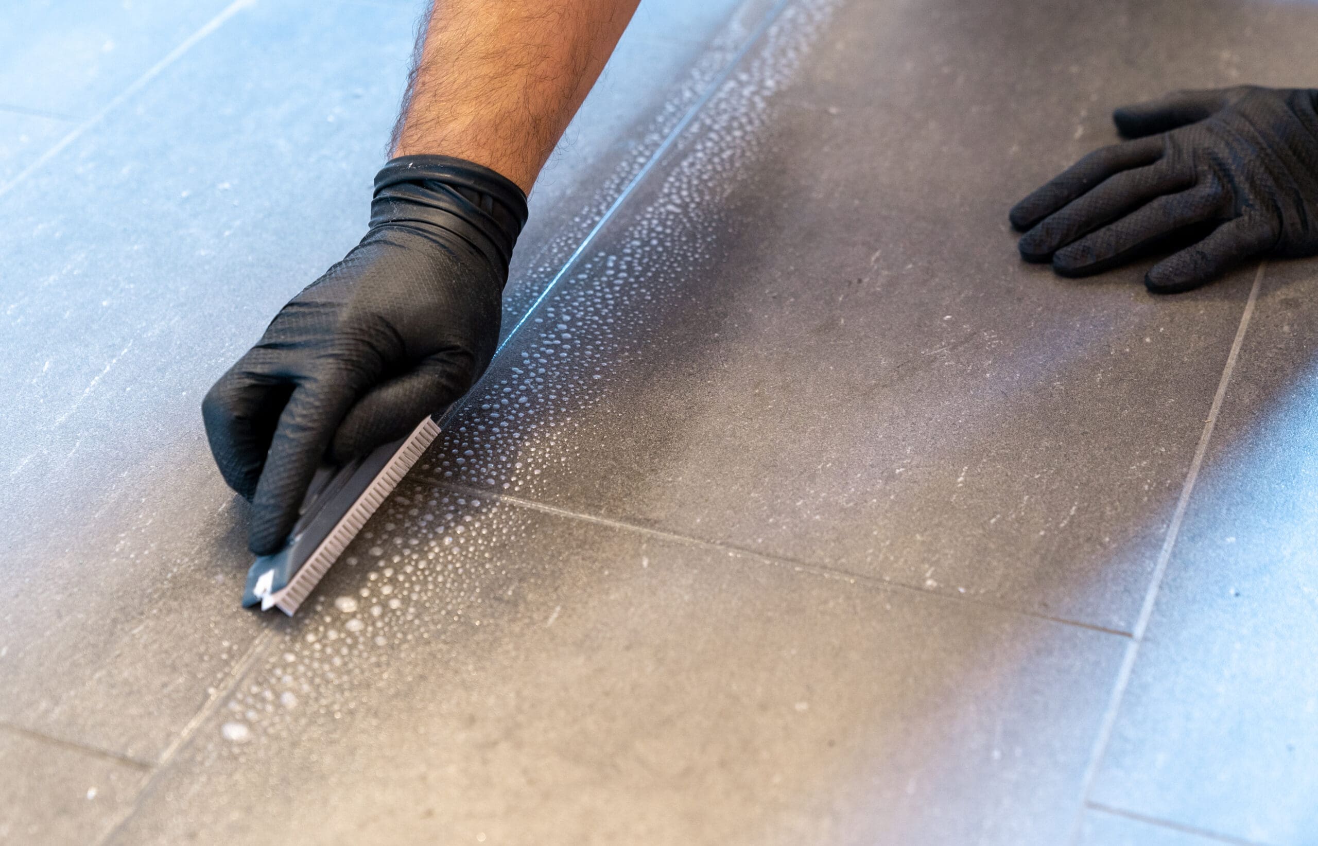 Close up of professional cleaner cleaning grout with a brush blade and foamy soap on a grey tiled bathroom floor